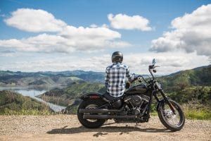 Man sitting on motorcycle, back to camera, looking at lake and mountains; image by Harley-Davidson via Unsplash.com.