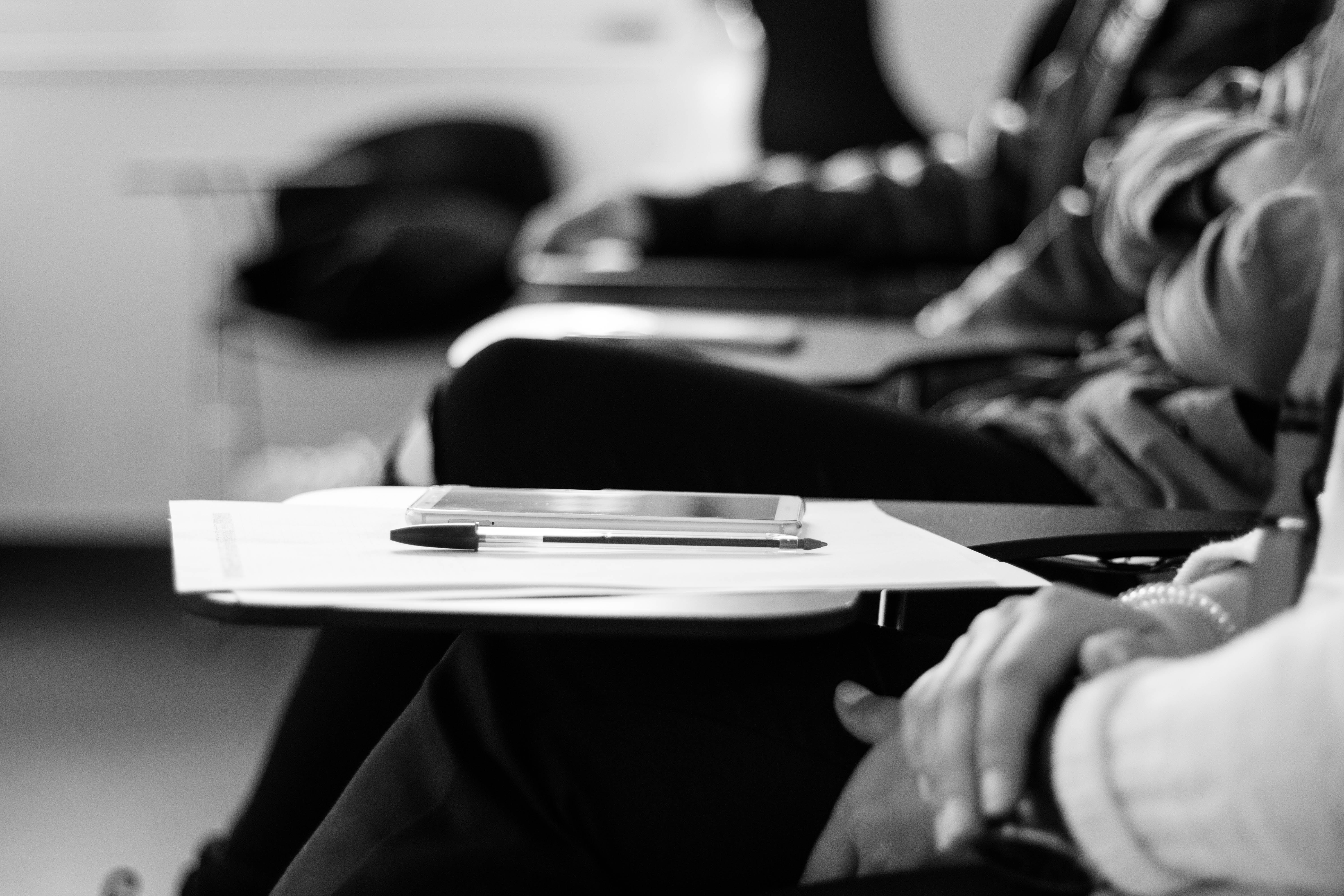 Grayscale photo of students sitting on chairs with papers and pens; image by Miguel Henriques, via Unsplash.com.