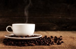 White ceramic mug and saucer with coffee beans on brown textile; image by Mike Kenneally, via Unsplash.com.