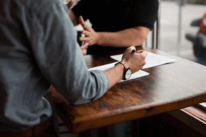 Two men reviewing paperwork at brown wooden table; image by Nik MacMillan, via Unsplash.com.