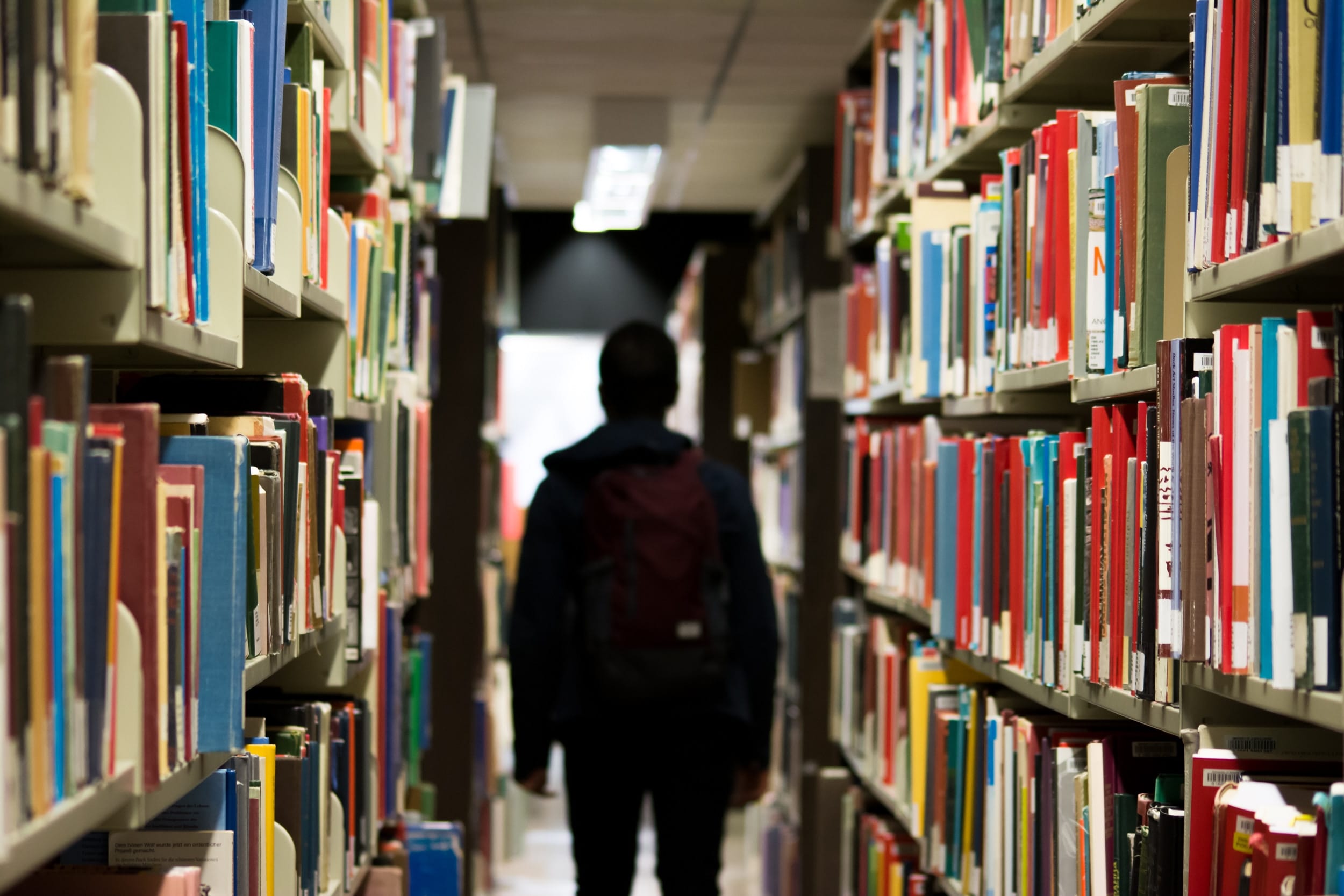 Male student with backpack walking between library shelves; image by Bantersnaps, via Unsplash.com.