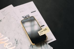 Black smartphone on top of papers. Phone has a sticker on the screen saying “Sign here.” Image by Kelly Sikkema, via Unsplash.com.