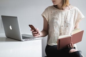 Woman holding book and smartphone looking at laptop; image by Burst, via Pexels.com.