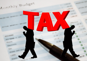 Budget paper and pen with silhouettes of two men walking away from each other and the word “Tax” in bold red letters; image by Geralt, via Pixabay.com.