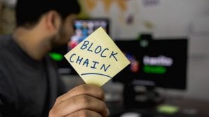 Man holding sticky note with “Blockchain” written on it; image by Hitesh Choudhary, via Unsplash.com.
