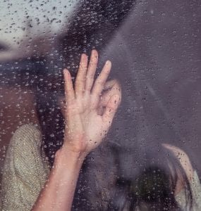 Woman touching raindrop covered window pane; image by Milada Vigerova, via Unsplash.com.