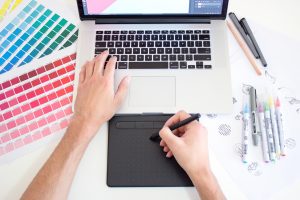 Graphic designer working on a Macbook laptop using a trackpad, color charts and markers; image by Theme Photos, via Unsplash.com.