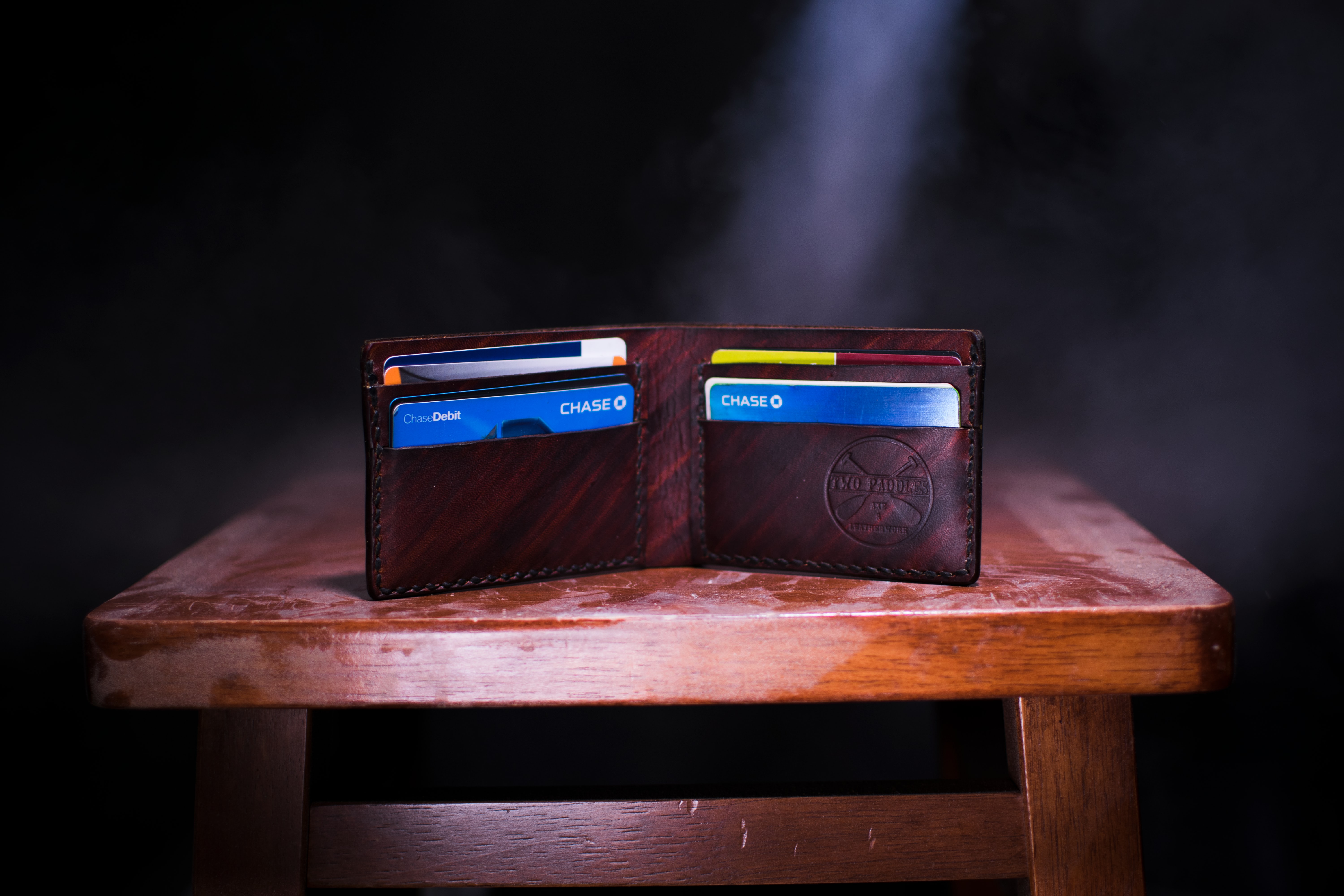 Handmade brown leather wallet with credit cards sitting on brown wooden stool; image by Two Paddles Axe and Leatherwork, via Unsplash.com.