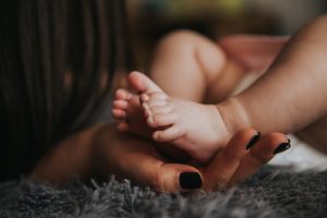 Infant's feet being held by a woman's hand with painted and manicured hands resting on a gray blanket; image by Alex Paserelu, via Unsplash.com.