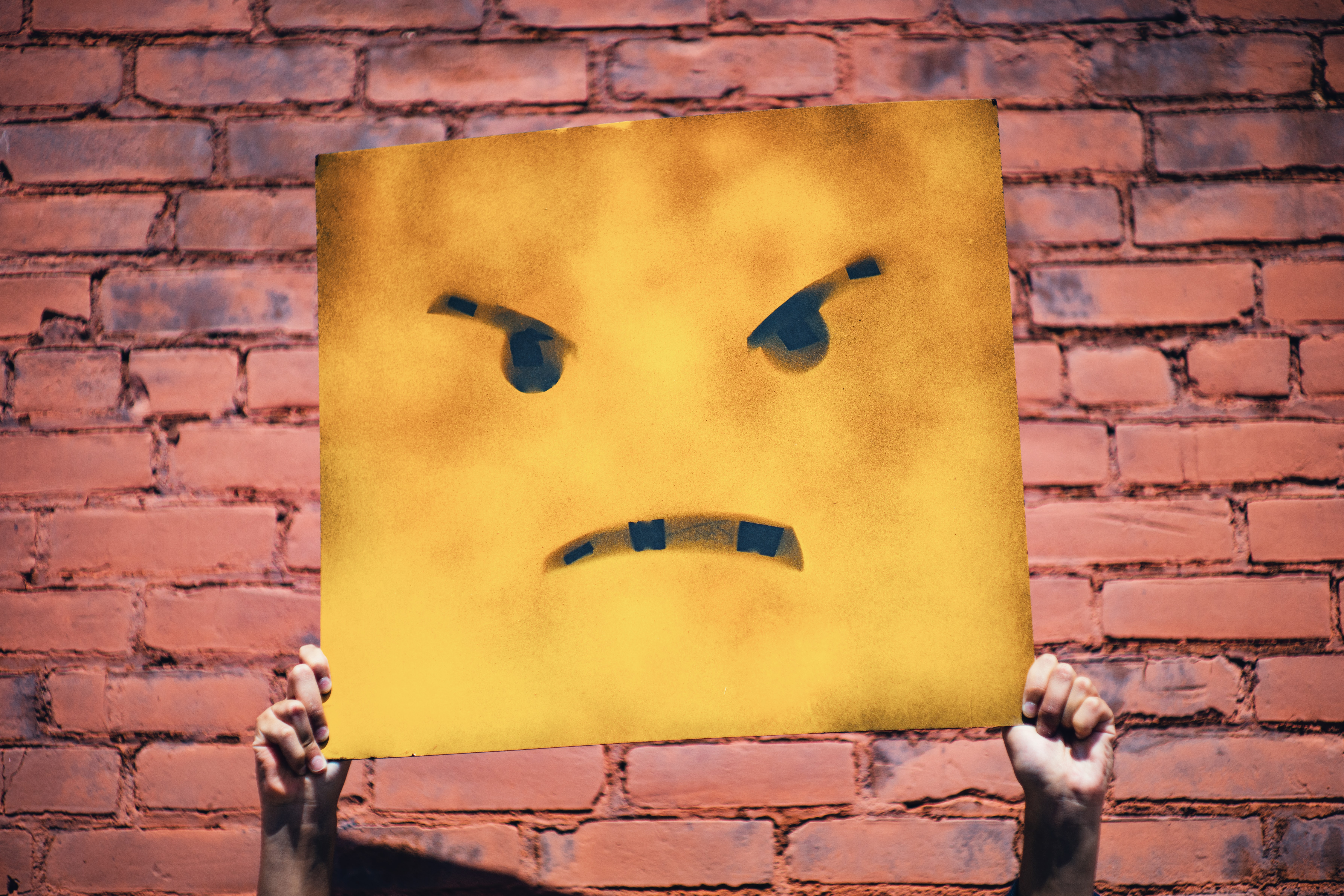 Person holding up sign with angry face on it standing in front of red brick wall; image by Andre Hunter, via Unsplash.com.