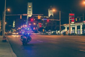 Police motorcycle with lights on parked at side of road at night; image by JP Valery, via Unsplash.com.