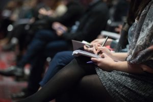 Woman writing in notebook with pen, seated in full auditorium; image by The Climate Reality Project, via Unsplash.com.