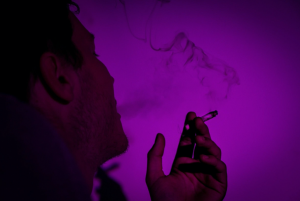 Profile view of man smoking in purple light; image by GRAS GRÜN, via Unsplash.com.