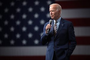 An older, white-haired man in a suit stands in front of an American flag background.