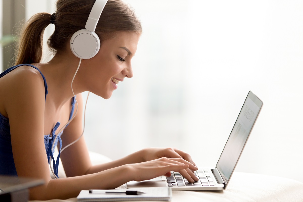 Side view portrait of smiling young woman in headphones typing on laptop; image by yanalya, via Freepik.com.
