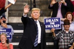 President Trump raising his hand at a rally, backed by fans waving signs.