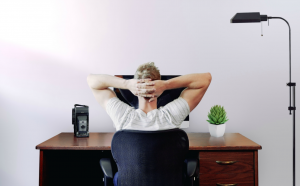 Man sitting at desk with hands clasped behind his head; image by Jason Strull, via Unsplash.com.