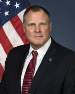 A white man in a suit positioned in front of an American flag.