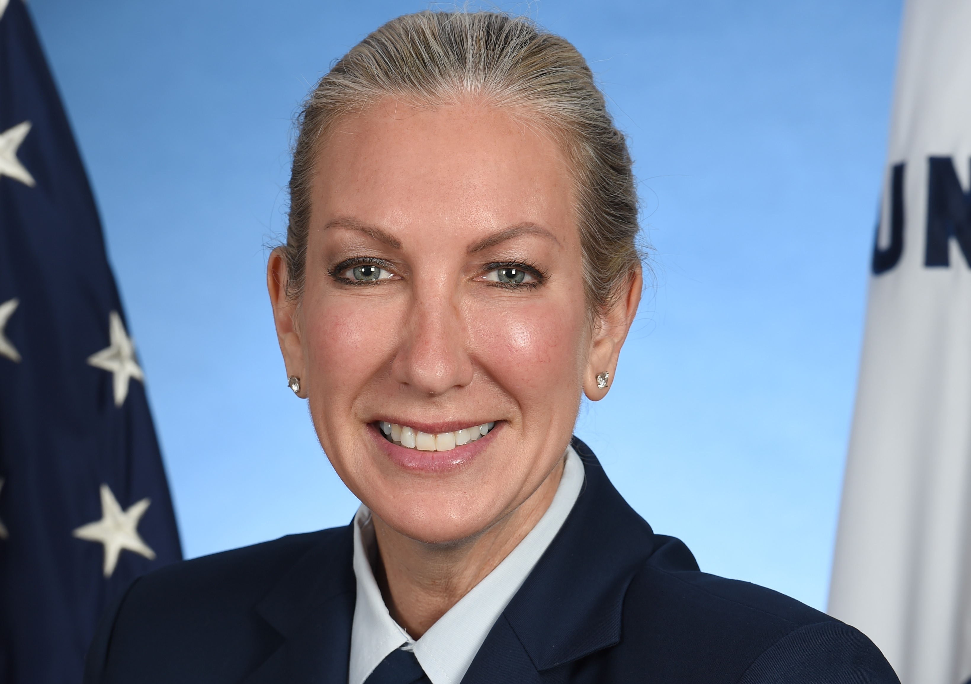 Portrait of a smiling woman in a military uniform, flanked by the flags of the United States and the Coast Guard.