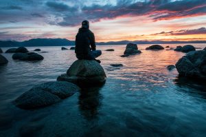 Man sitting on rock in body of water; image by Keegan Houser, via Unsplash.com.