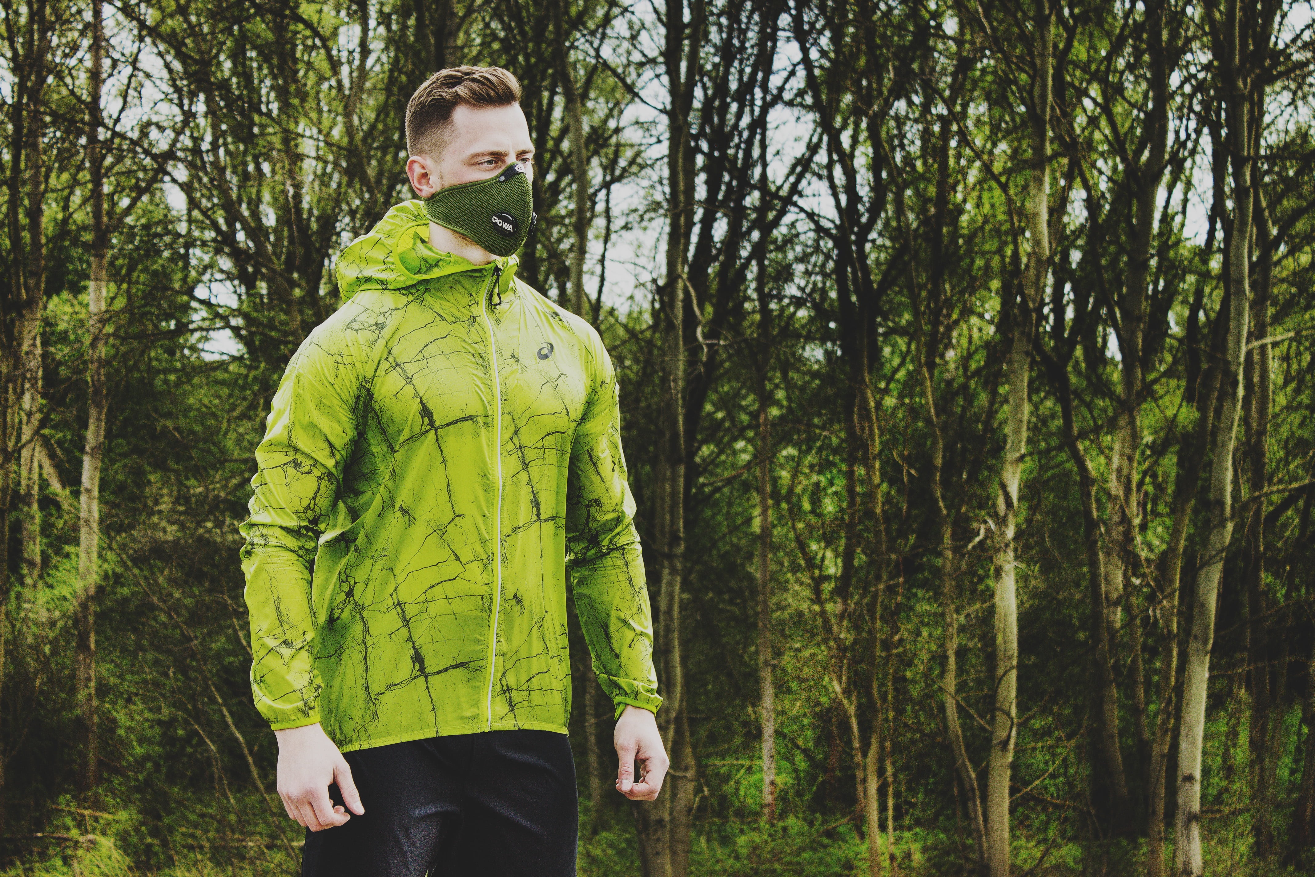 Man wearing green hoodie and mask beside trees; image via Pikrepo.com.