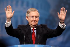 A greying, older man in a dark suit, at a podium, with his hands raised.