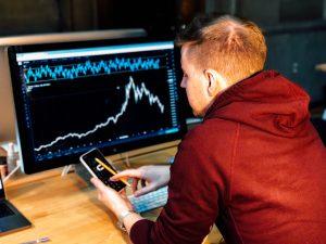 Man with laptop and phone working on investing; image by Chris Liverani, via Unsplash.com.