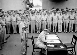 General Douglas MacArthur signs as Supreme Allied Commander during formal surrender ceremonies on the USS MISSOURI in Tokyo Bay. Behind General MacArthur are Lieutenant General Jonathan Wainwright and Lieutenant General A. E. Percival. Image by U.S. Navy, Wikimedia Commons, public domain.