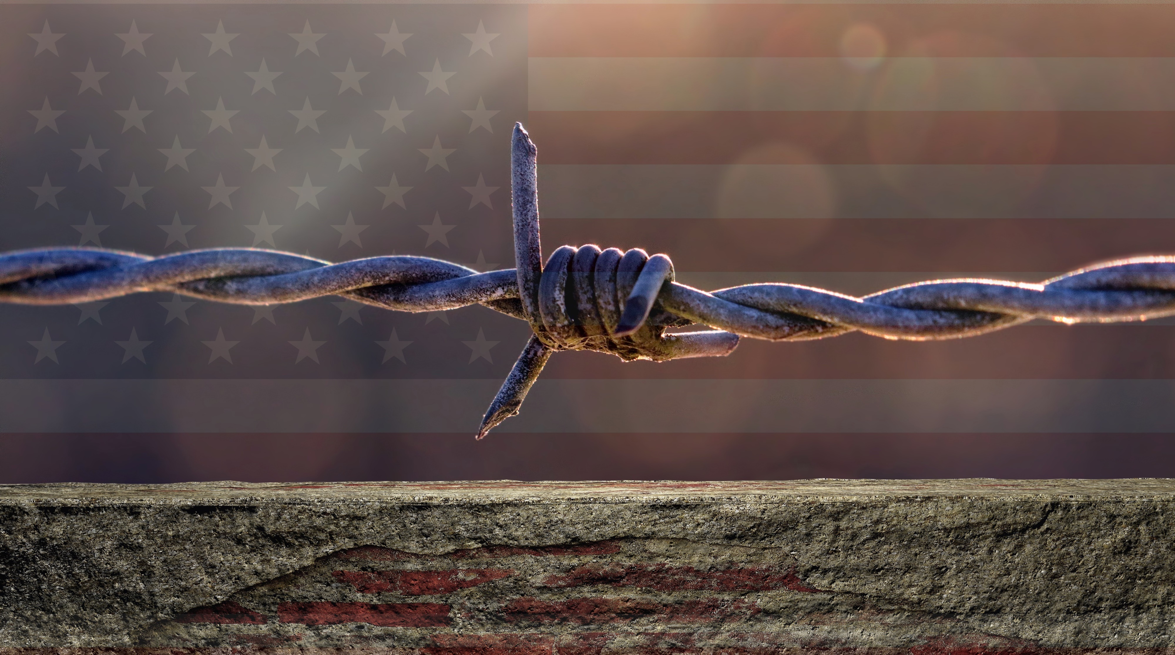 A wall topped with a strand of barbed wire, with a shadowy American flag in the distant background.