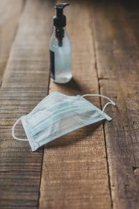 Hand sanitizer and mask sitting on wooden table; image by Anshu A., via Unsplash.com.