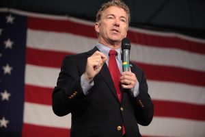 A man in a suit speaks into a microphone in front of a large American flag.