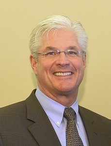 A white-haired white man, grinning and wearing a suit.