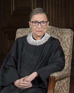 Ruth Bader Ginsburg, wearing her judicial robe and trademark collar, sitting in a chair.