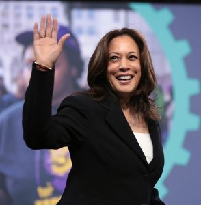 A woman in a dark blazer waves towards the camera.