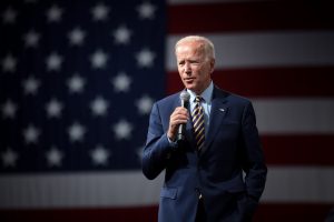 A man in a suit speaking into a microphone in front of an American flag backdrop.