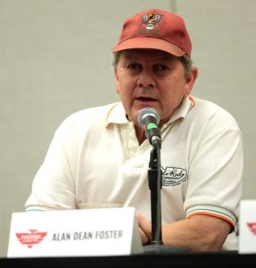 A man in a white polo shirt and faded red hat sits at a table at a convention.