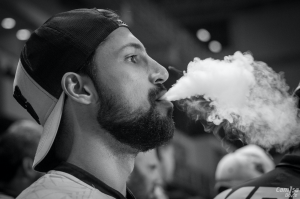 Grayscale photo of man in backwards baseball cap vaping; image by Miguel Arcanjo Saddi, via Pexels.com.