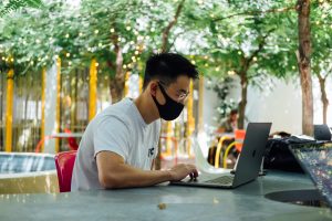 Man in mask working on laptop at outdoor table; image by Paul Hanaoka, via Unsplash.com.