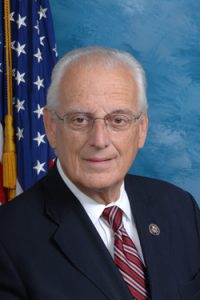 Portrait of a white-haired man in a suit and tie, in front of the American flag.