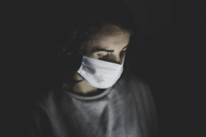 Man in crewneck shirt wearing mask, looking down at smartphone; image by engin akyurt, via Unsplash.com.