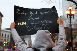 Person in grey hoodie holding sign saying, “Girls Just Wanna Have FUNdamental Rights”; image by Lucia, via Unsplash.com.