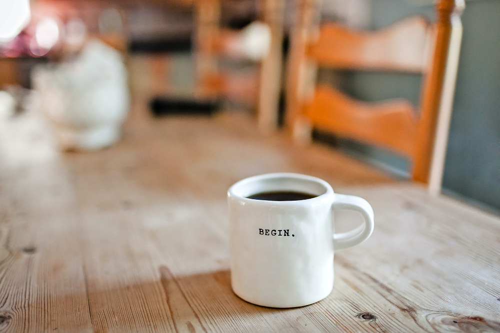 White mug with “Begin” written on it, sitting on a wooden table; image by Danielle MacInnes, via Unsplash.com.