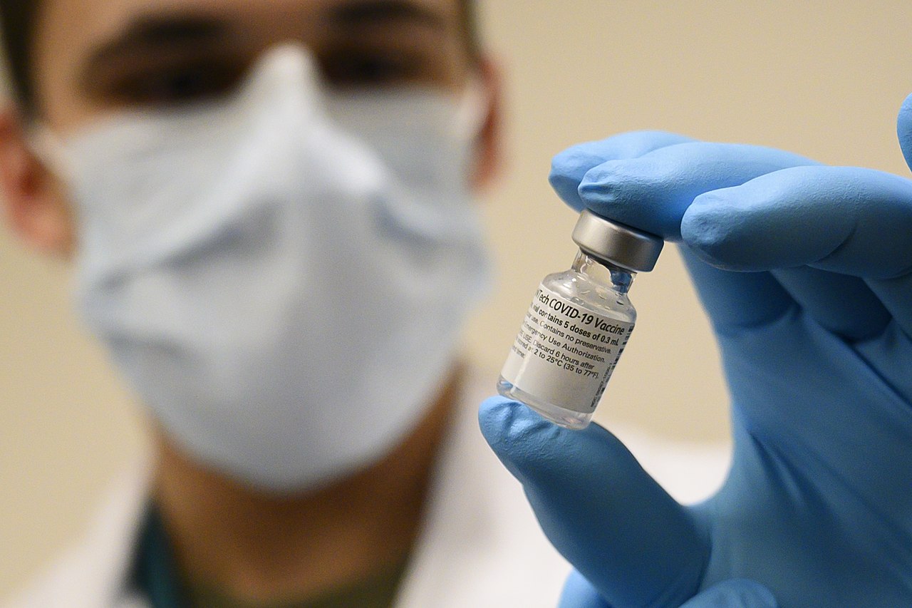A blurred masked individual with a blue medical glove holds a vial of COVID vaccine.