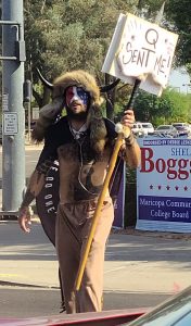 Jacob Chansely in his iconic political costume, including face paint and horned headdress, carrying a sign that says "Q Sent Me".