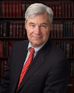 A silver-haired man in a suit with a red tie smiles for the camera.