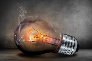 A broken incandescent lightbulb glows dimly against a grey background, emitting a wisp of smoke.