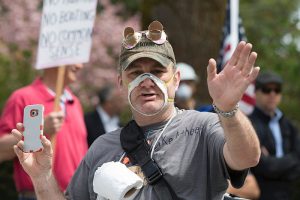 An anti-mask protester, wearing a mask with the center cut out, his mouth and nose plainly visible, and a roll of toilet paper hanging from a string around his neck.