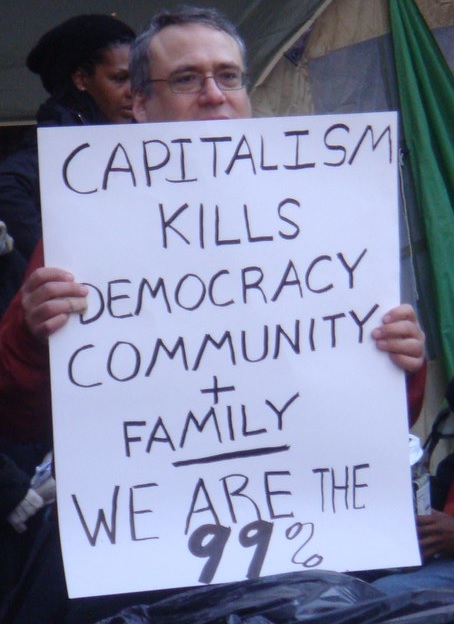 A man holds a sign that reads, "Capitalism Kills Democracy Community + Family - We are the 99%"
