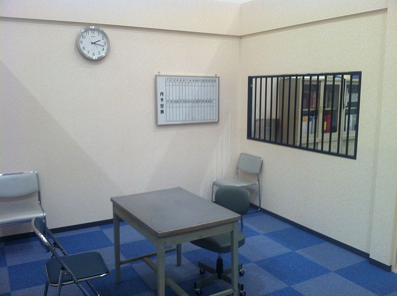 A sparsely furnished room with a tile floor, small table, uncomfortable chairs, a wall clock, and bars on the window.