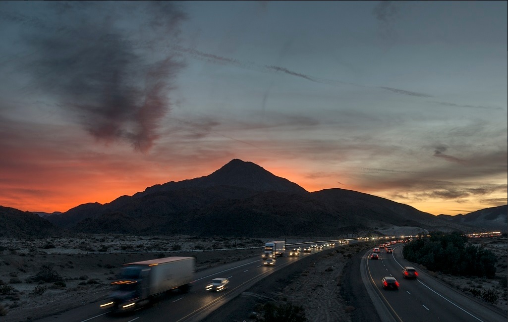 Cars and semi-trucks speed along a freeway at sunset.